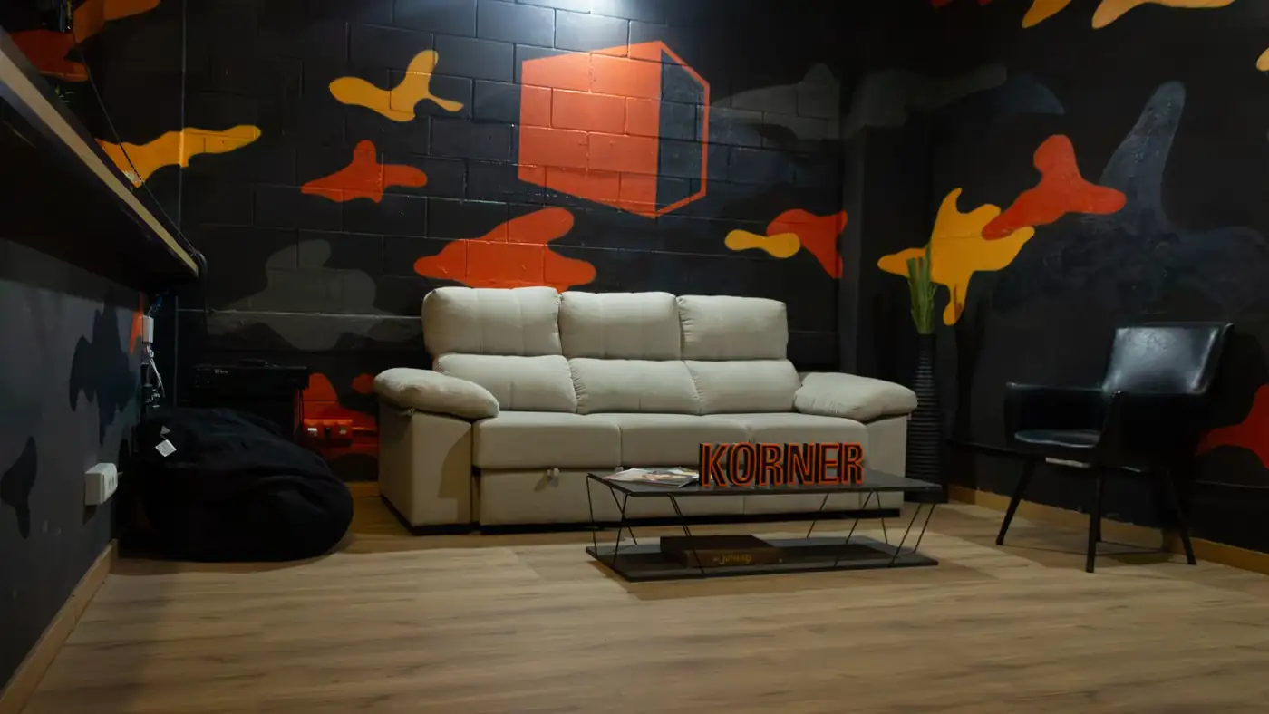 Zona de estar de arriba: sofá, mesa con el letrero de Korner y la pared de camuflaje con el isotipo